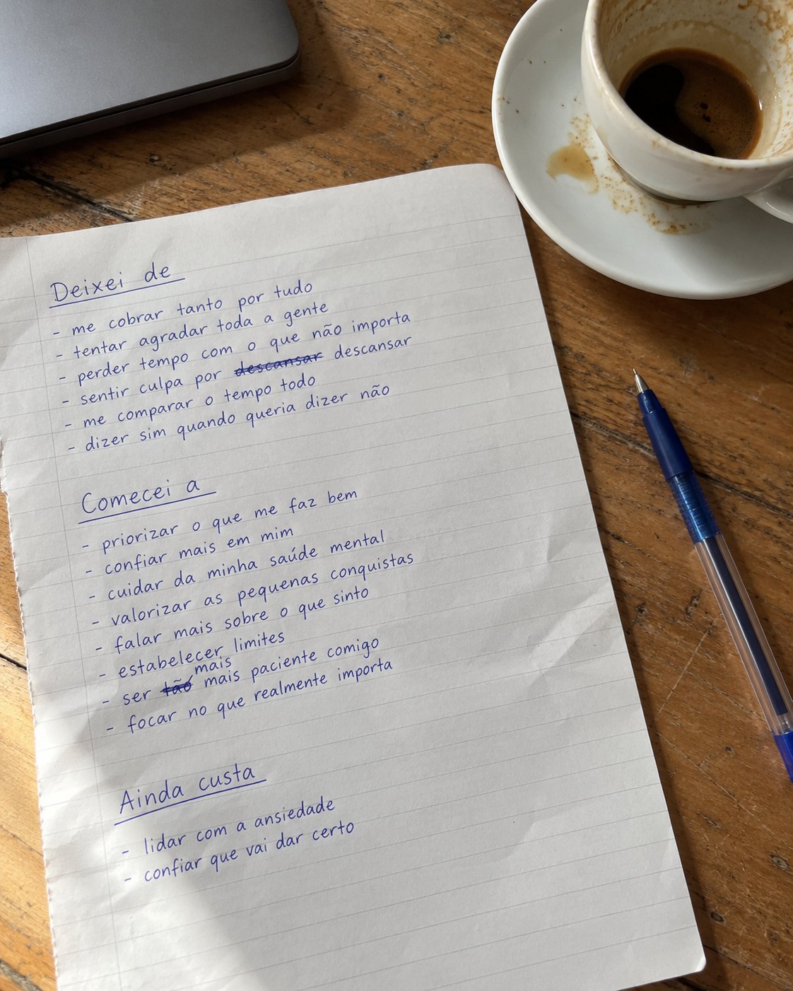 Folha de caderno escrita à mão sobre uma mesa de madeira, com as listas «Deixei de», «Comecei a» e «Ainda custa», ao lado de uma chávena de café vazia e uma caneta.
