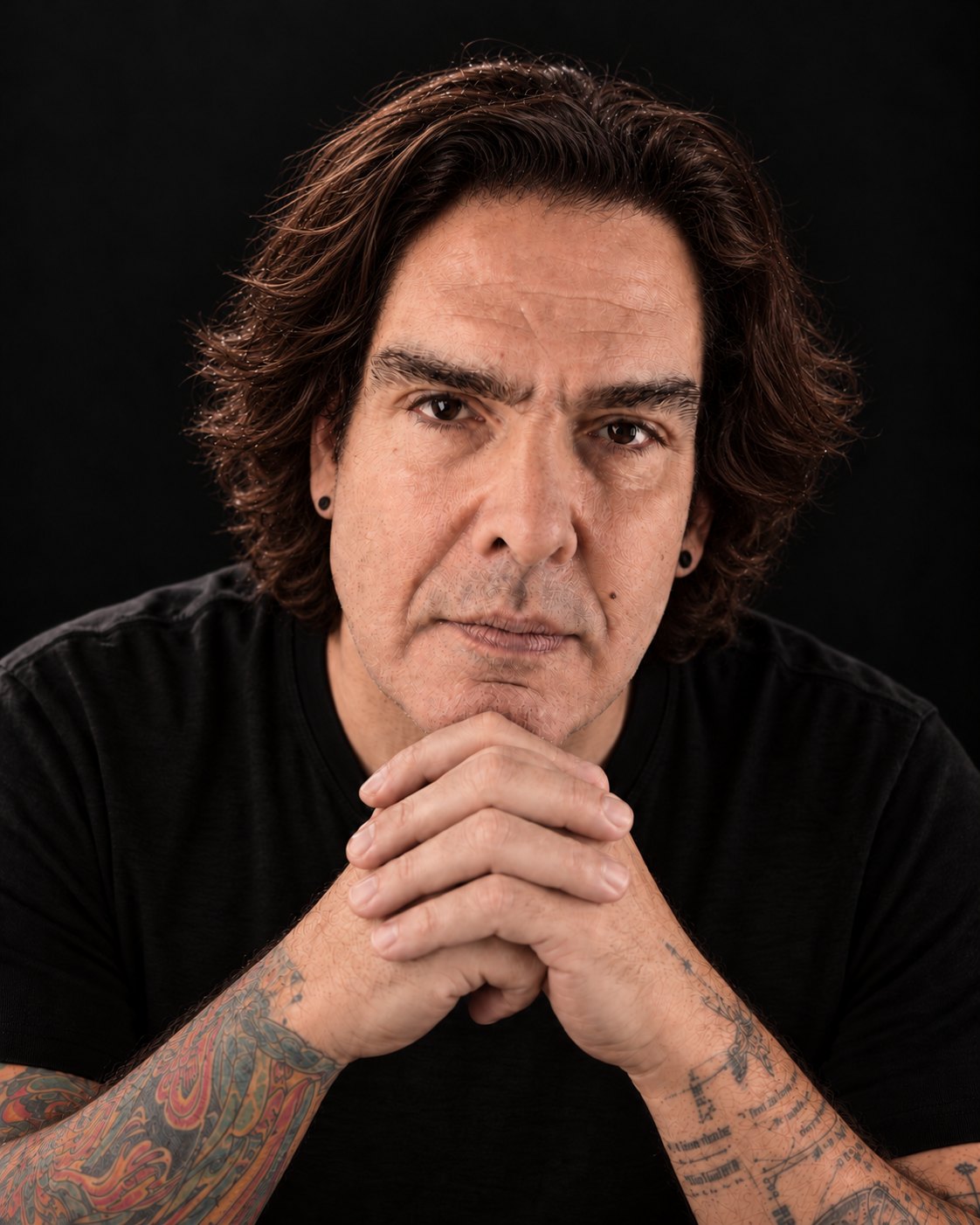 João Costta de frente, de t-shirt preta, com as mãos entrelaçadas junto ao queixo e os antebraços tatuados, sobre fundo escuro.