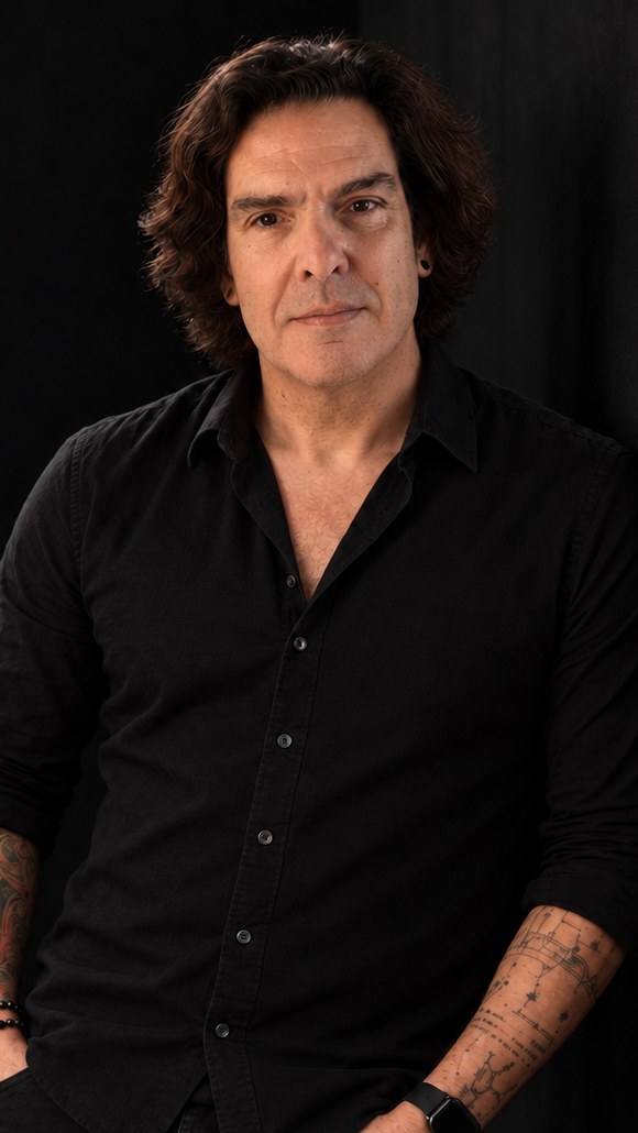 João Costta, de camisa preta e braços tatuados, encostado a uma parede escura.