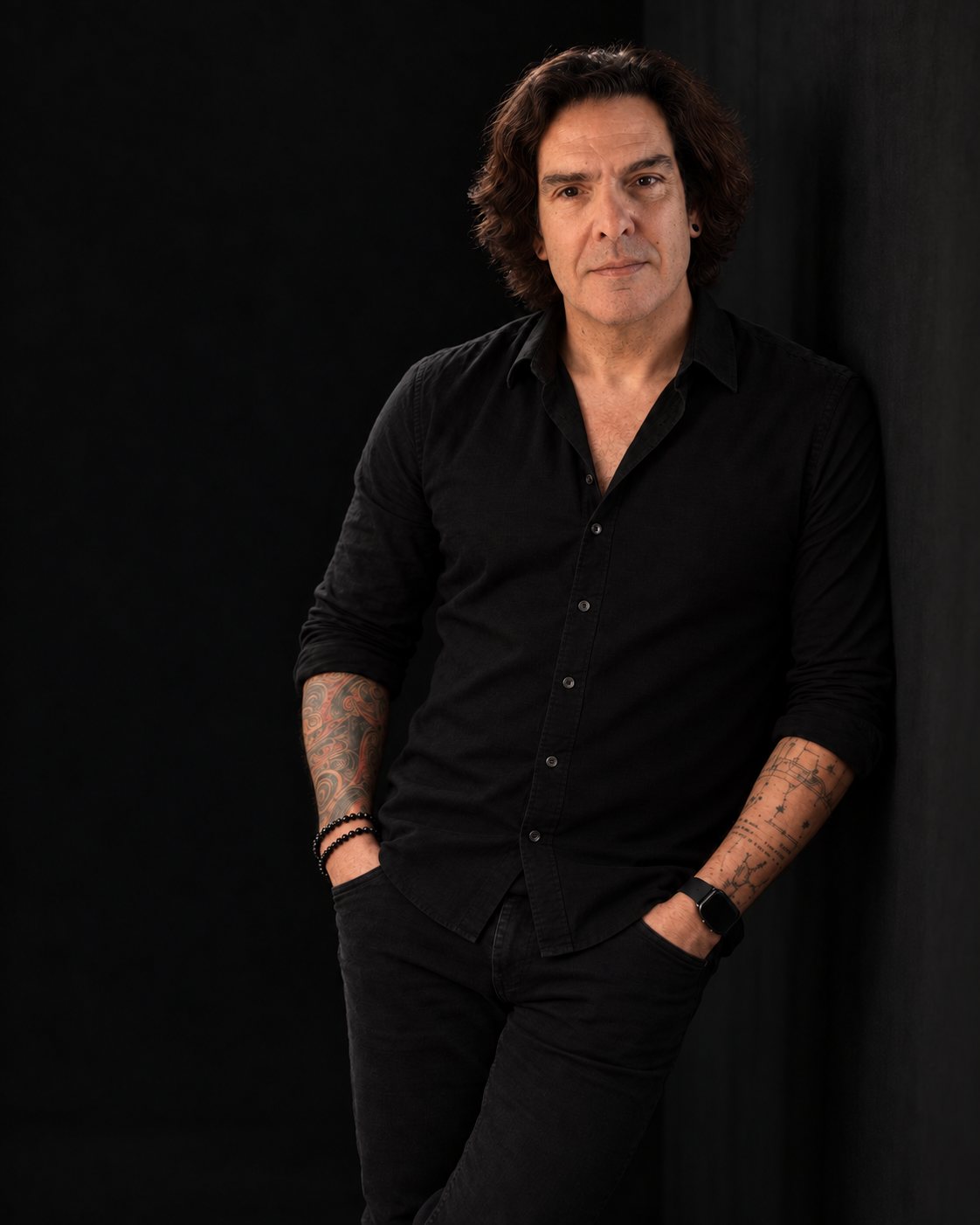 João Costta, de pé, encostado a uma parede, de camisa e calças pretas, sobre fundo escuro.
