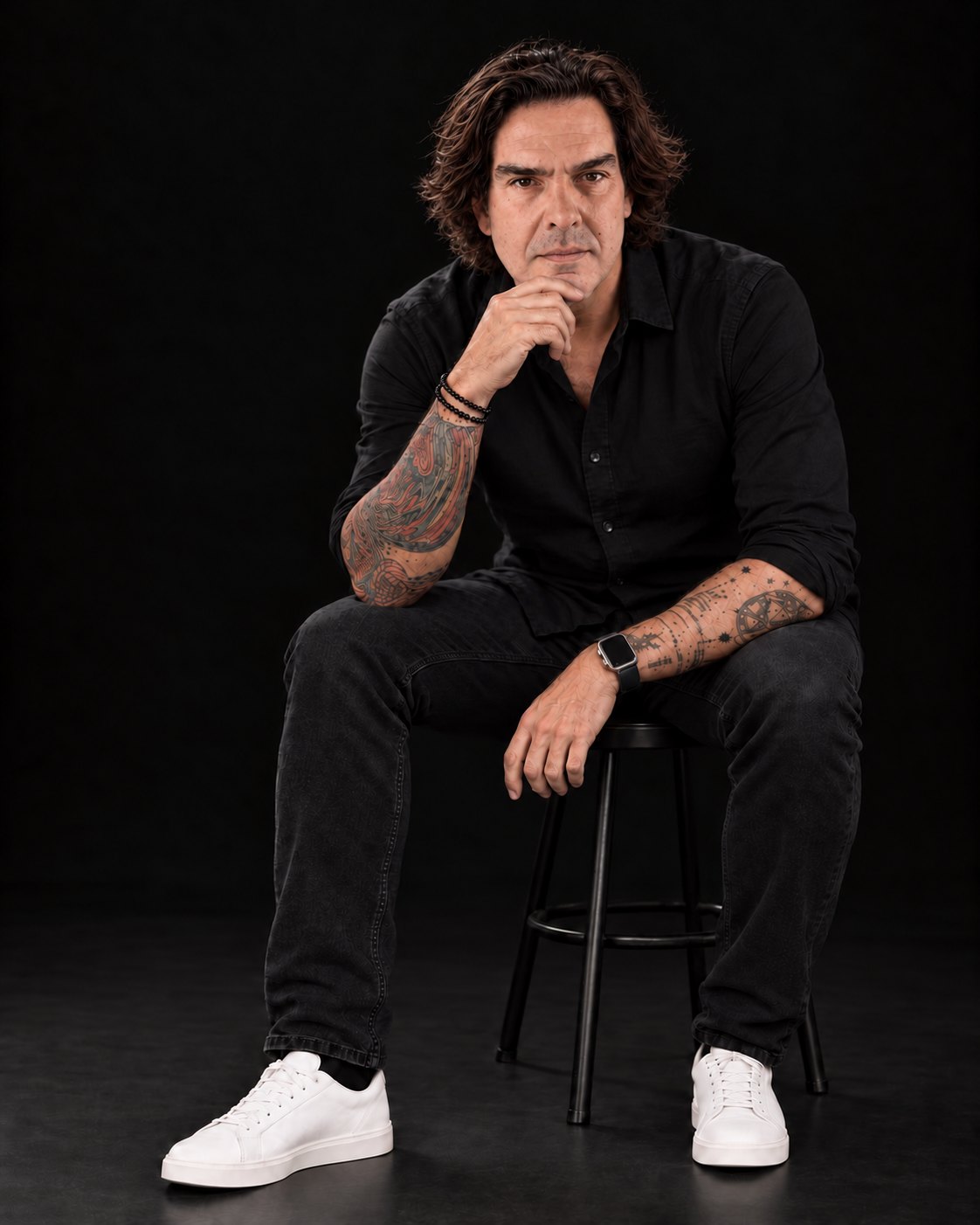 João Costta sentado num banco alto, de camisa preta e ténis brancos, com a mão no queixo, sobre fundo escuro.